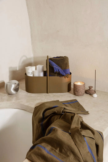 A bathroom scene features a beige counter with a brown caddy, toiletries, rolled towels, a lit candle, an incense holder, and the ferm LIVING Komo Mini Vases Set of 3. A blue-striped towel is draped over the bathtub’s edge.