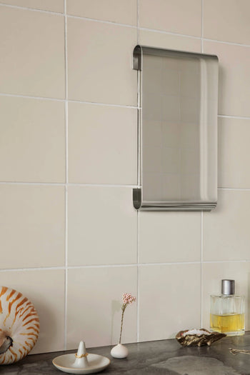 A small, rectangular metal-framed mirror hangs on a tiled bathroom wall above a countertop with a seashell, the ferm LIVING Komo Mini Vases (set of 3), one holding a flower, and a bottle of perfume.