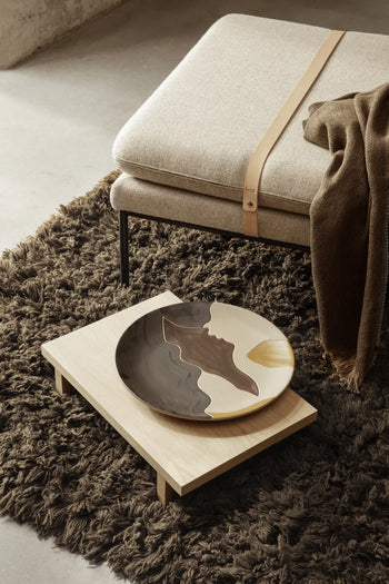 A modern living space showcases a shaggy brown rug, a ferm LIVING Kona Side Table crafted from FSC-certified oak veneer, a decorative ceramic plate with abstract patterns, and a beige cushioned bench highlighted by a leather strap and brown throw blanket.