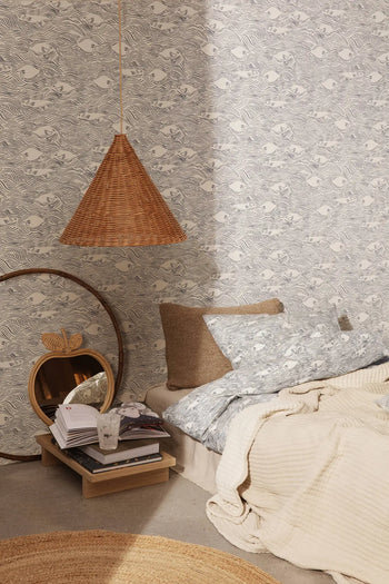 A cozy bedroom corner features a beige bed with patterned bedding, textured cushions, and a wicker pendant lamp. A round mirror hangs above the ferm LIVING Kona Side Table by ferm LIVING, styled with books and water against gray animal print wallpaper.