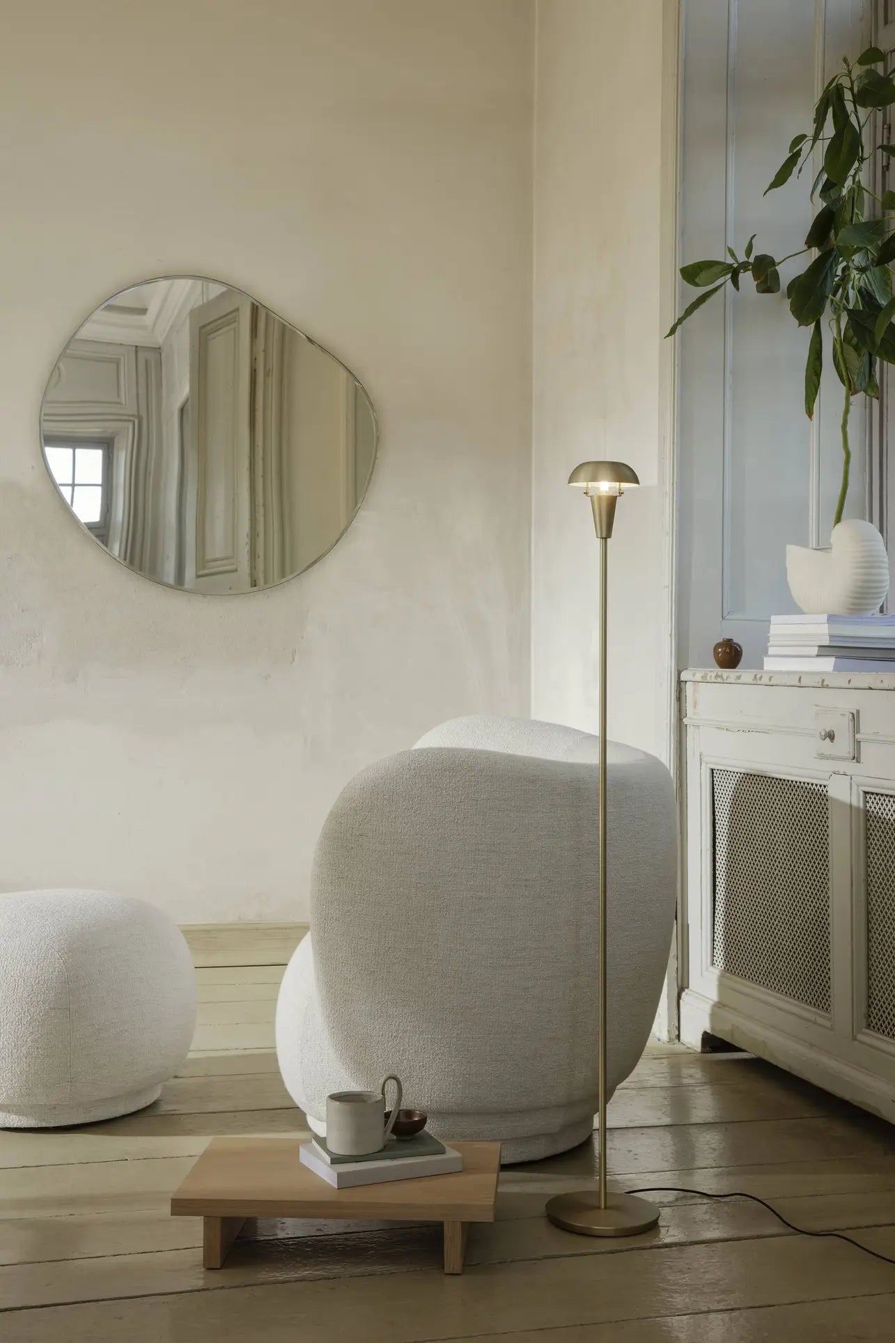 A cozy, modern living room with a round white armchair, ottoman, gold floor lamp, ferm LIVING Kona Side Table in FSC-certified oak veneer, potted plant, abstract wall mirror, and a low wooden coffee table with a cup and teapot.