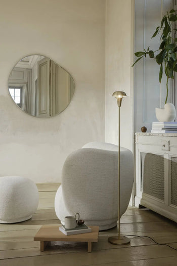 A cozy, modern living room with a round white armchair, ottoman, gold floor lamp, ferm LIVING Kona Side Table in FSC-certified oak veneer, potted plant, abstract wall mirror, and a low wooden coffee table with a cup and teapot.