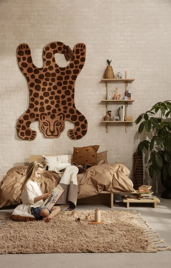 A child sits reading on a beige rug in a cozy bedroom featuring tan bedding, Japanese-inspired decor, wooden shelves, toys, a potted plant, and the ferm LIVING Kona Side Table. A large leopard wall hanging completes the look.