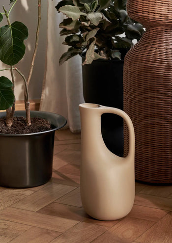 The ferm LIVING Liba Watering Can in modern beige with a curved handle sits on a wooden floor next to a black recycled plastic plant pot with greenery and a large woven basket, all bathed in natural light.
