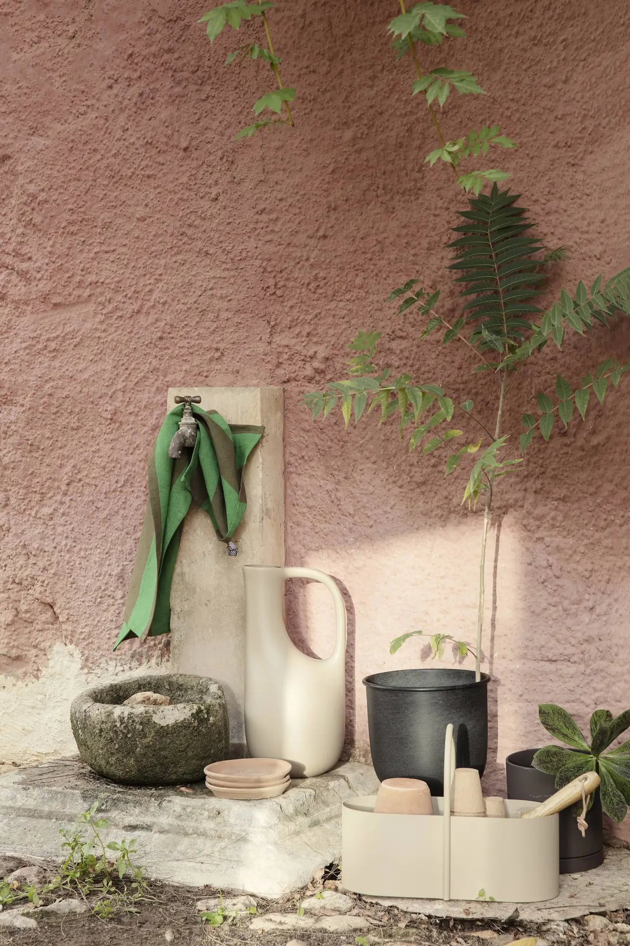 A rustic outdoor scene features a stone basin, beige pitcher, bowls, potted plants, the ferm LIVING Liba Watering Can, and a green cloth hanging from a wall-mounted faucet on a textured pink wall with leafy branches overhead.