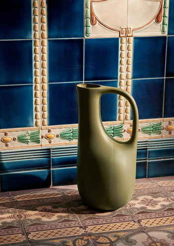 The ferm LIVING Liba Watering Can, by ferm LIVING, in tall matte olive-green ceramic with a large handle, rests on patterned tiles against a decorative wall of blue and beige geometric and floral tile designs.
