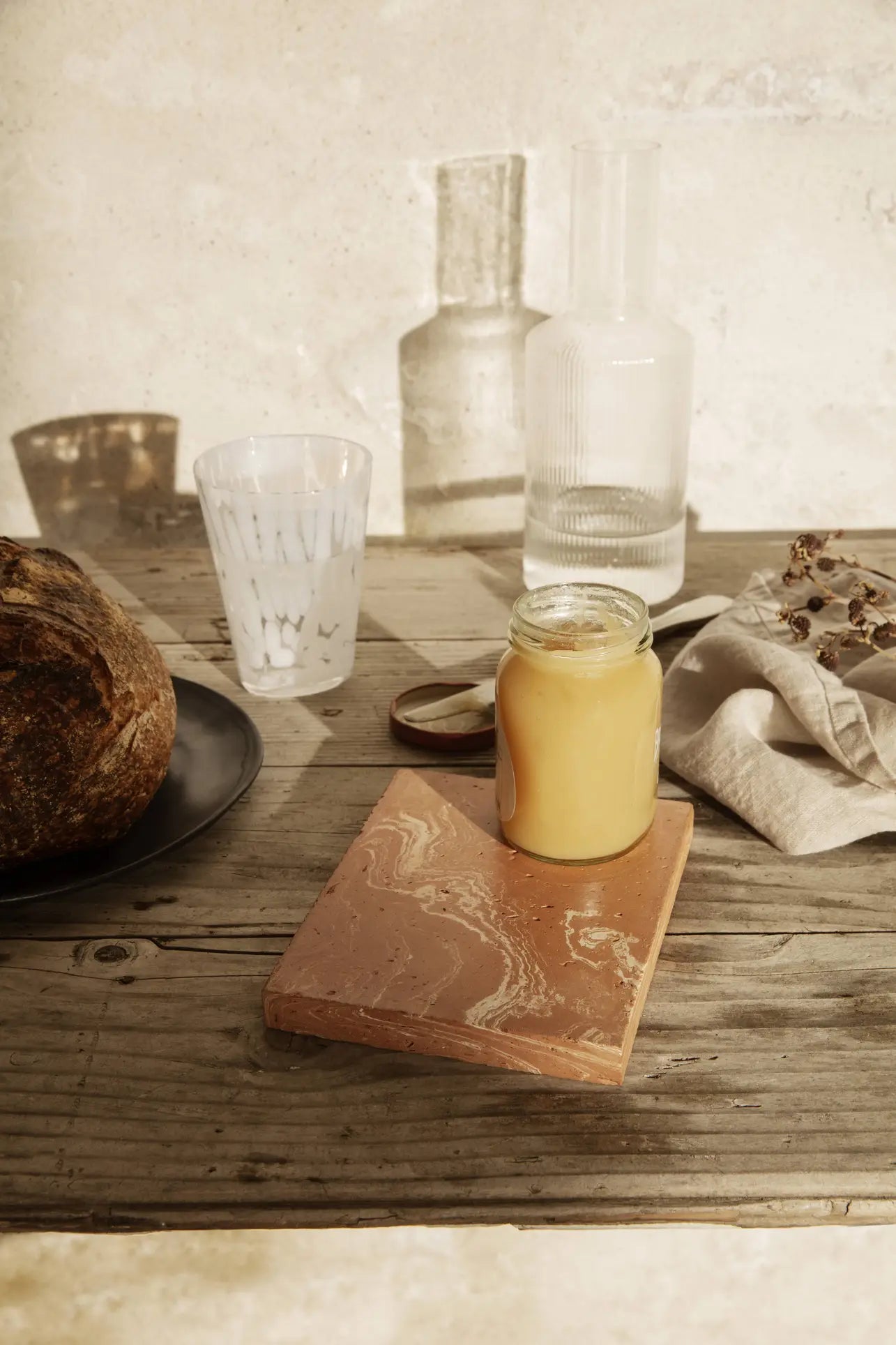 A jar of yellow spread rests on a ferm LIVING Merge Trivet atop a rustic wooden table with bread, a white cup, a glass carafe, and a beige napkin decorated with dried flowers. Sunlight creates gentle shadows.