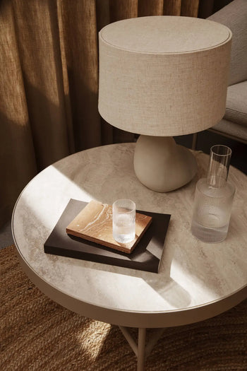 A round marble-top table with a beige lamp, clear ribbed glass, matching carafe, brown book, black notebook, and a handmade ferm LIVING Merge Trivet by ferm LIVING sits on a woven rug with sunlight streaming across the surface.