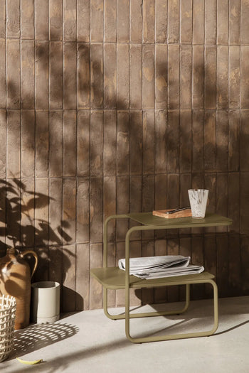 A compact olive-green side table with two shelves displays a glass, folded cloth, book, and the handmade ferm LIVING Merge Trivet, set by a textured brown tiled wall with sunlight casting plant shadows nearby.