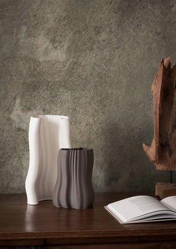 The ferm LIVING Moire Vase, Small sits with another ribbed ceramic vase on a wooden surface beside an open book and abstract wood sculpture, highlighting the artistry of 3D-printed clay against a textured gray wall backdrop.