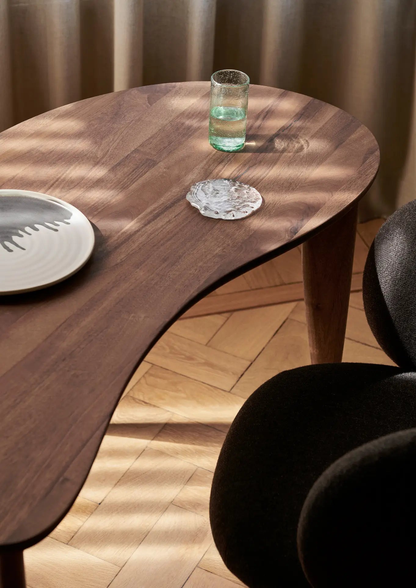 A kidney-shaped wooden coffee table displays a ferm LIVING Momento Glass Stones S and a white textured coaster on a tray. Sunlight creates shadows, while a black chair and parquet floor add to the rooms sophisticated ambiance.