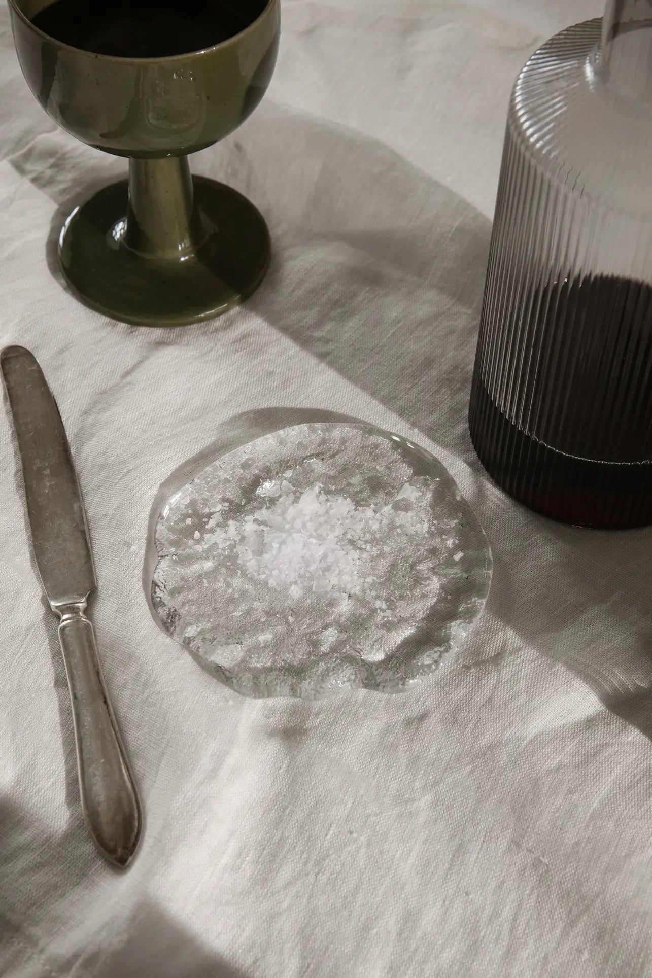 A glass plate with flaky salt sits on a white tablecloth, enhanced by ferm LIVING Momento Glass Stones S, Set of 2, alongside a vintage-style knife, green goblet, and ribbed bottle filled with dark liquid.