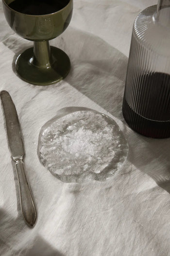 A glass plate with flaky salt sits on a white tablecloth, enhanced by ferm LIVING Momento Glass Stones S, Set of 2, alongside a vintage-style knife, green goblet, and ribbed bottle filled with dark liquid.