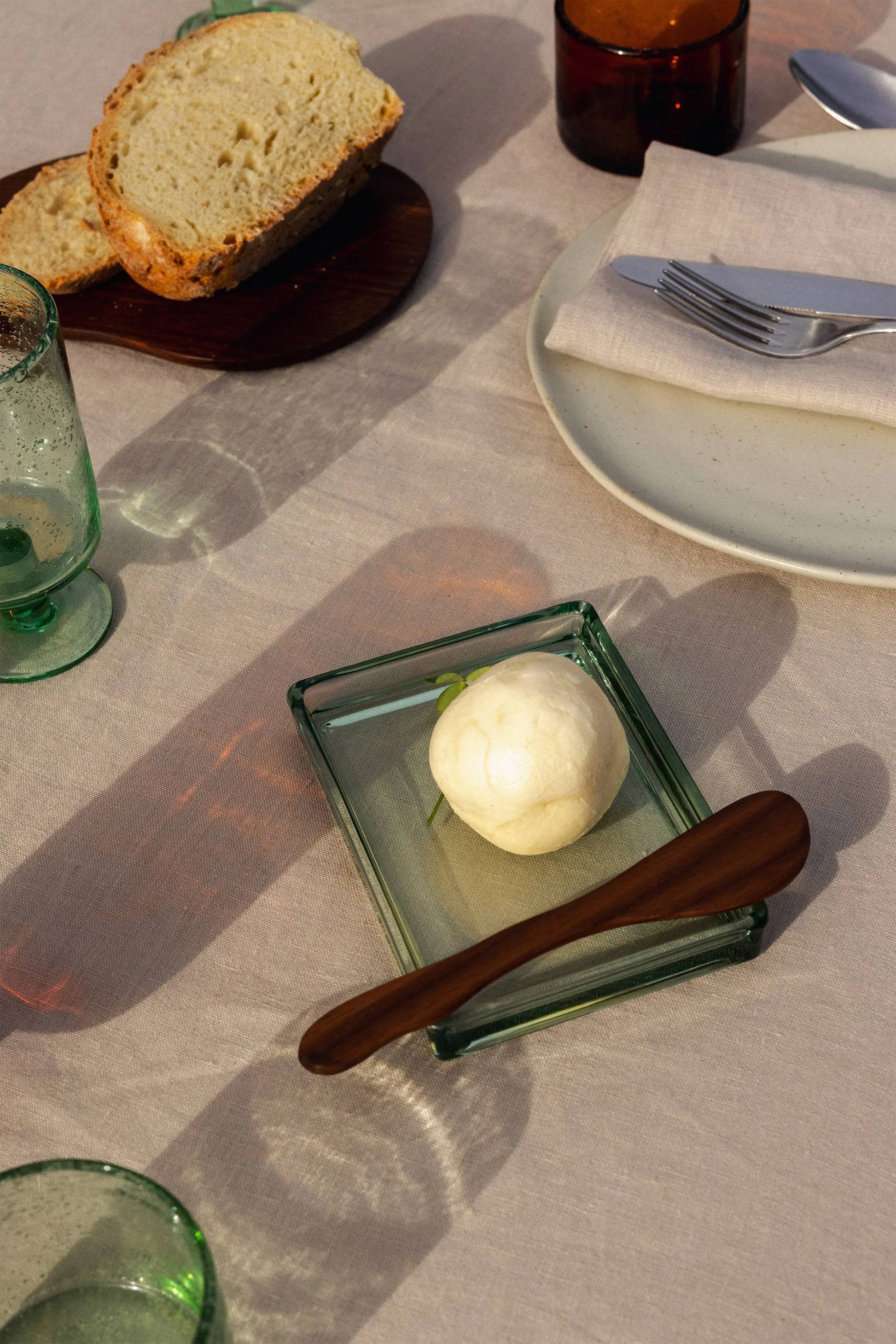 A table set with the ferm LIVING Oli Box—a recycled glass butter dish from ferm LIVING—holding a round pat of butter and wooden knife, plus green glassware, linen napkin on a plate, sliced bread, and a beige tablecloth.