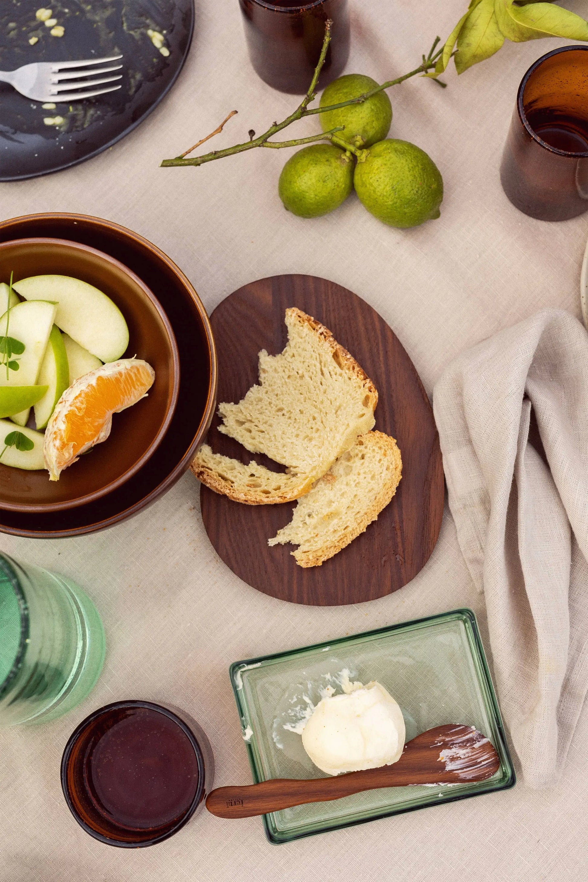 A table set with sliced bread, a bowl of fruit, brown cups, limes on a branch, a beige napkin on a light cloth, and the versatile ferm LIVING Oli Box—a recycled glass container by ferm LIVING—used for storing butter.