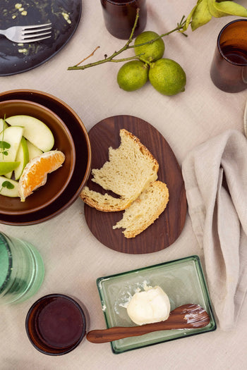 A table set with sliced bread, a bowl of fruit, brown cups, limes on a branch, a beige napkin on a light cloth, and the versatile ferm LIVING Oli Box—a recycled glass container by ferm LIVING—used for storing butter.