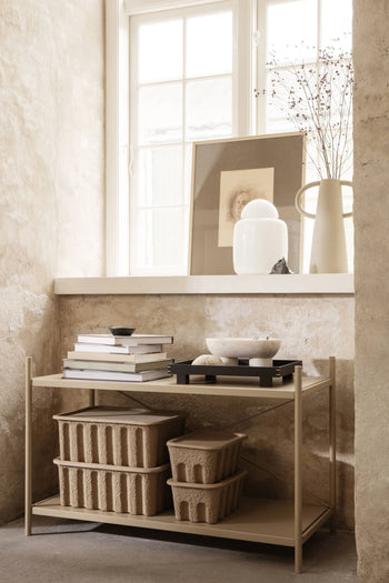 A minimalist beige shelf stands by a sunlit window, holding stacked books, decorative bowls, and a set of two ferm LIVING Paper Pulp Box Large. Above, a vase with dried branches and framed art rest on the windowsill against a textured wall.