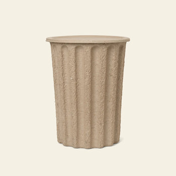 The ferm LIVING Paper Pulp Paper Bin features a cylindrical, beige, fluted design made from recycled paper pulp with a textured surface and flat top, echoing classical columns, set against a plain light background.