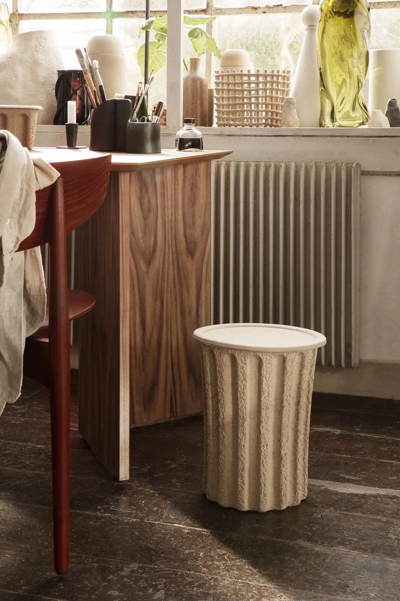 A cozy workspace features a wooden desk with art supplies stored in a ferm LIVING Paper Pulp Paper Bin, a red chair, textured beige stool, and a large window with plants and ceramics. Sunlight streams in, creating a warm, inviting atmosphere.