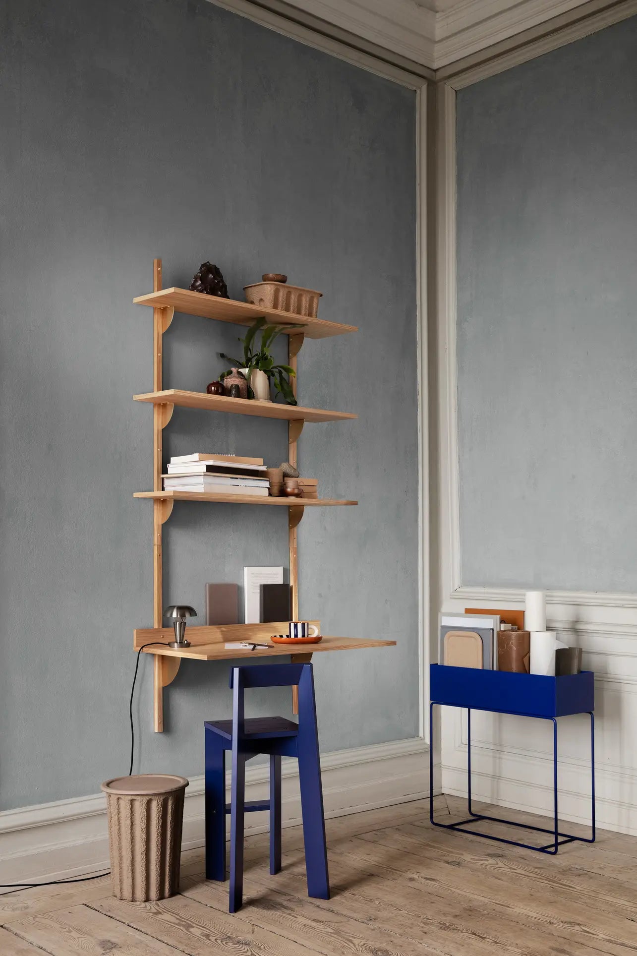 Minimalist workspace with wooden shelves, a small wooden desk, blue chair, and the ferm LIVING Paper Pulp Paper Bin by ferm LIVING for storage—plus a textured stool on light wood floors set against classic molding.