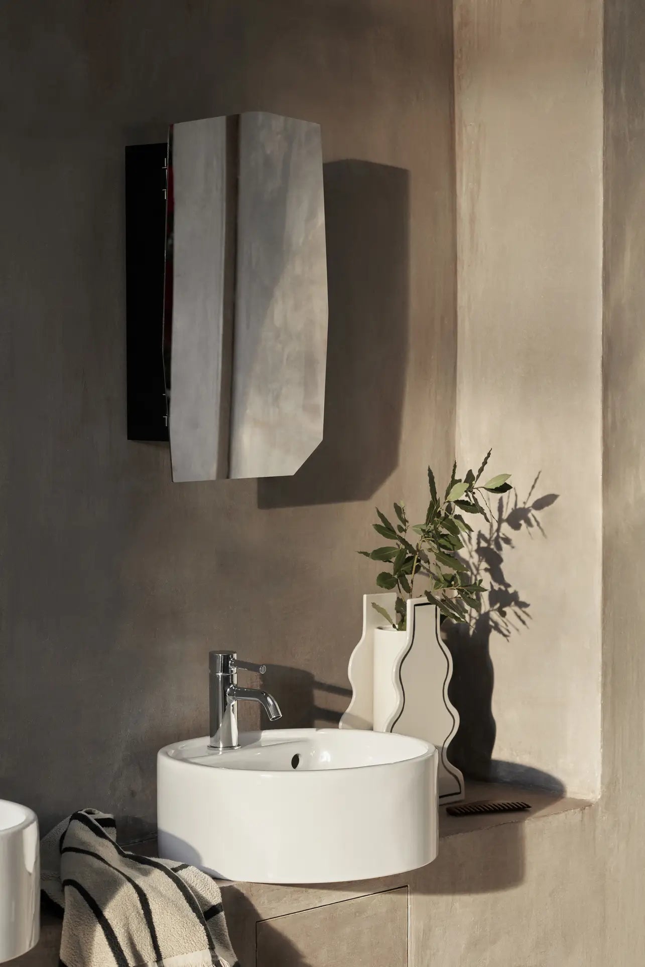 A modern bathroom features a round white sink with a silver faucet and two ferm LIVING Paste Vase, Curvy from ferm LIVING holding green branches. Above is an angular mirror on a beige textured wall with sunlight casting soft shadows.
