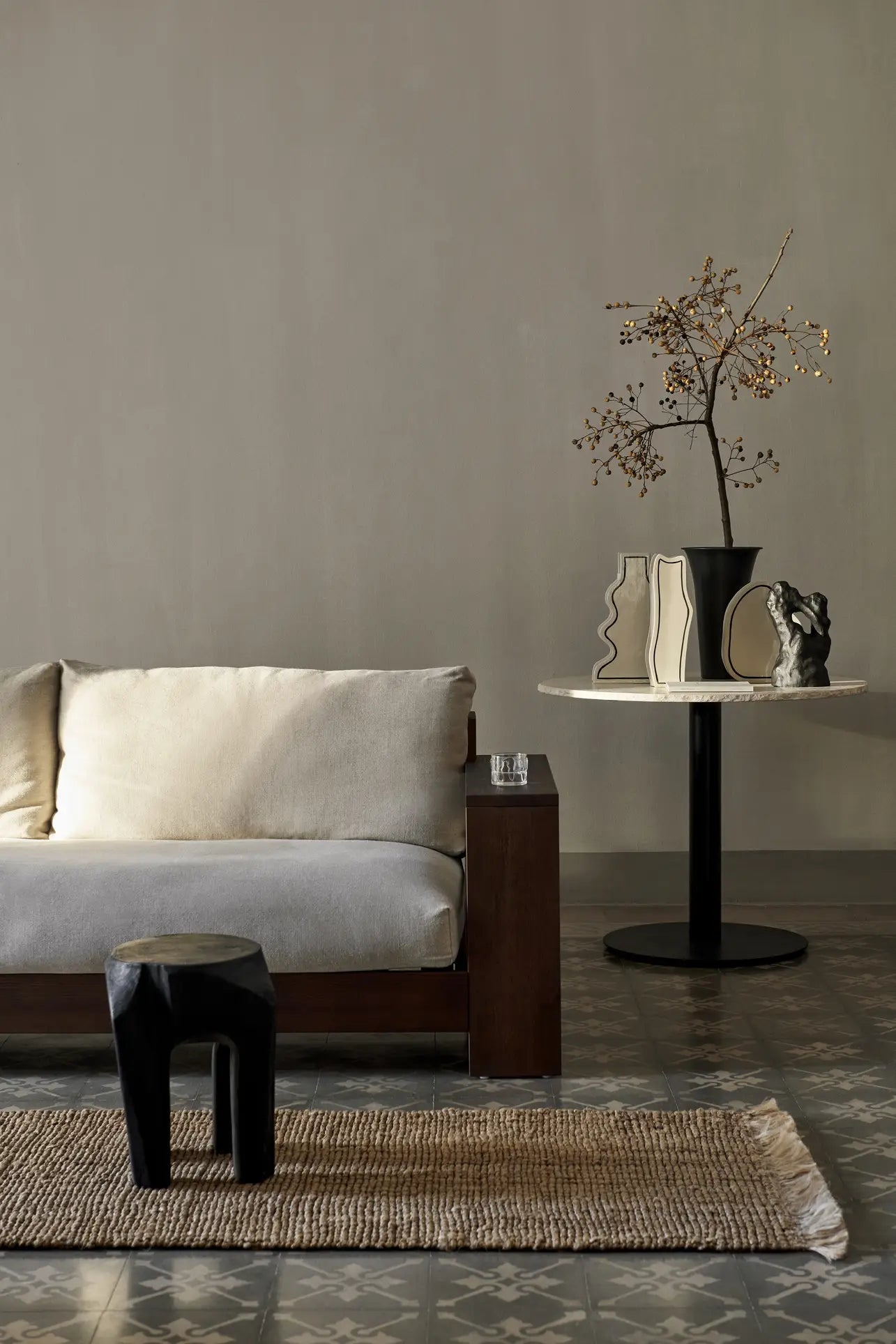 A minimalist living room featuring a beige sofa, black stool, round table with black base, abstract vases like the ferm LIVING Paste Vase Curvy, a black sculptural piece, bare branch in a vase, and ceramic sheets on patterned tile flooring.