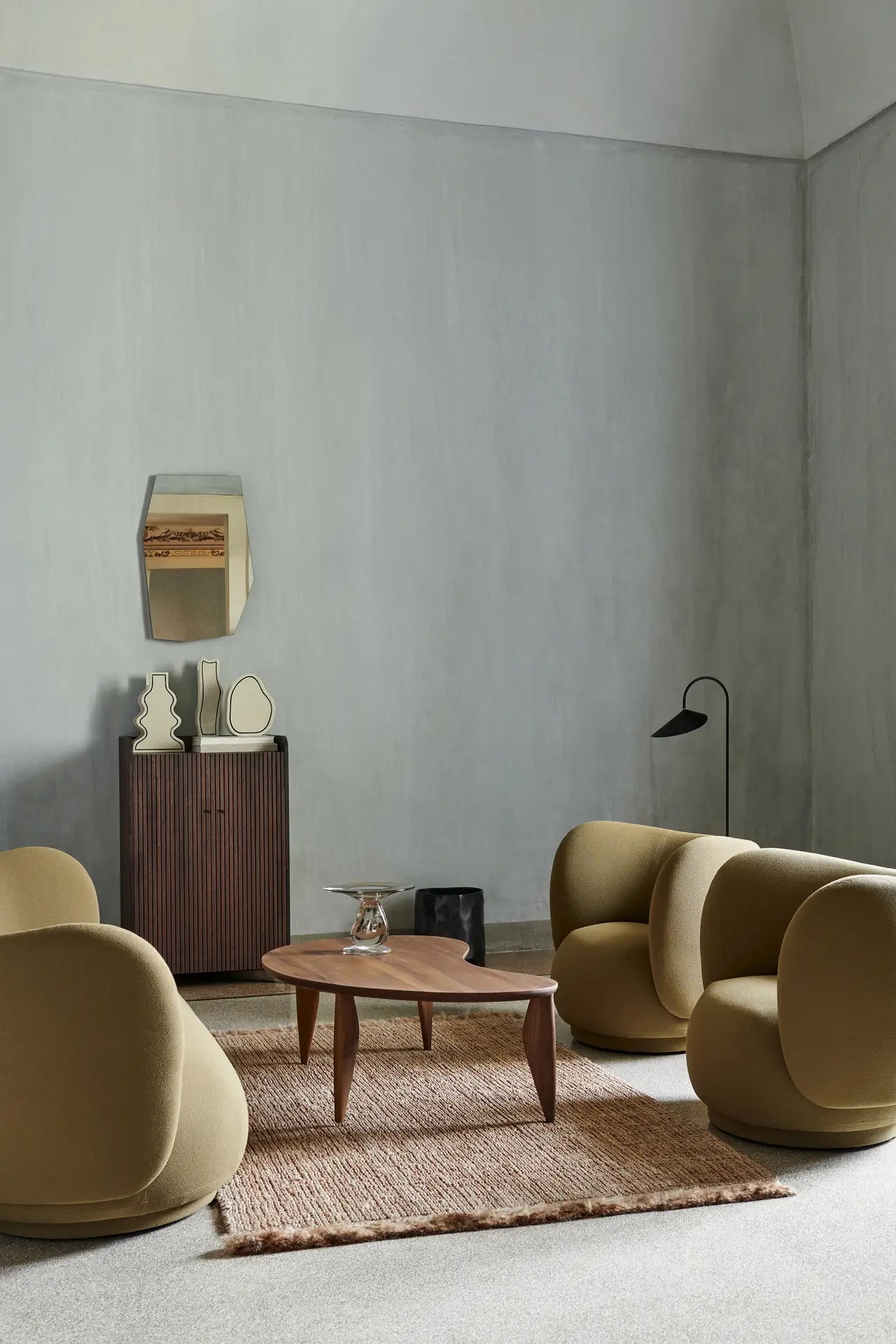 A modern living room features three curved mustard chairs, a wooden coffee table, and a textured rug on neutral carpet. The ferm LIVING Paste Vase, Curvy sits on the small cabinet beneath a geometric mirror on the pale gray wall.