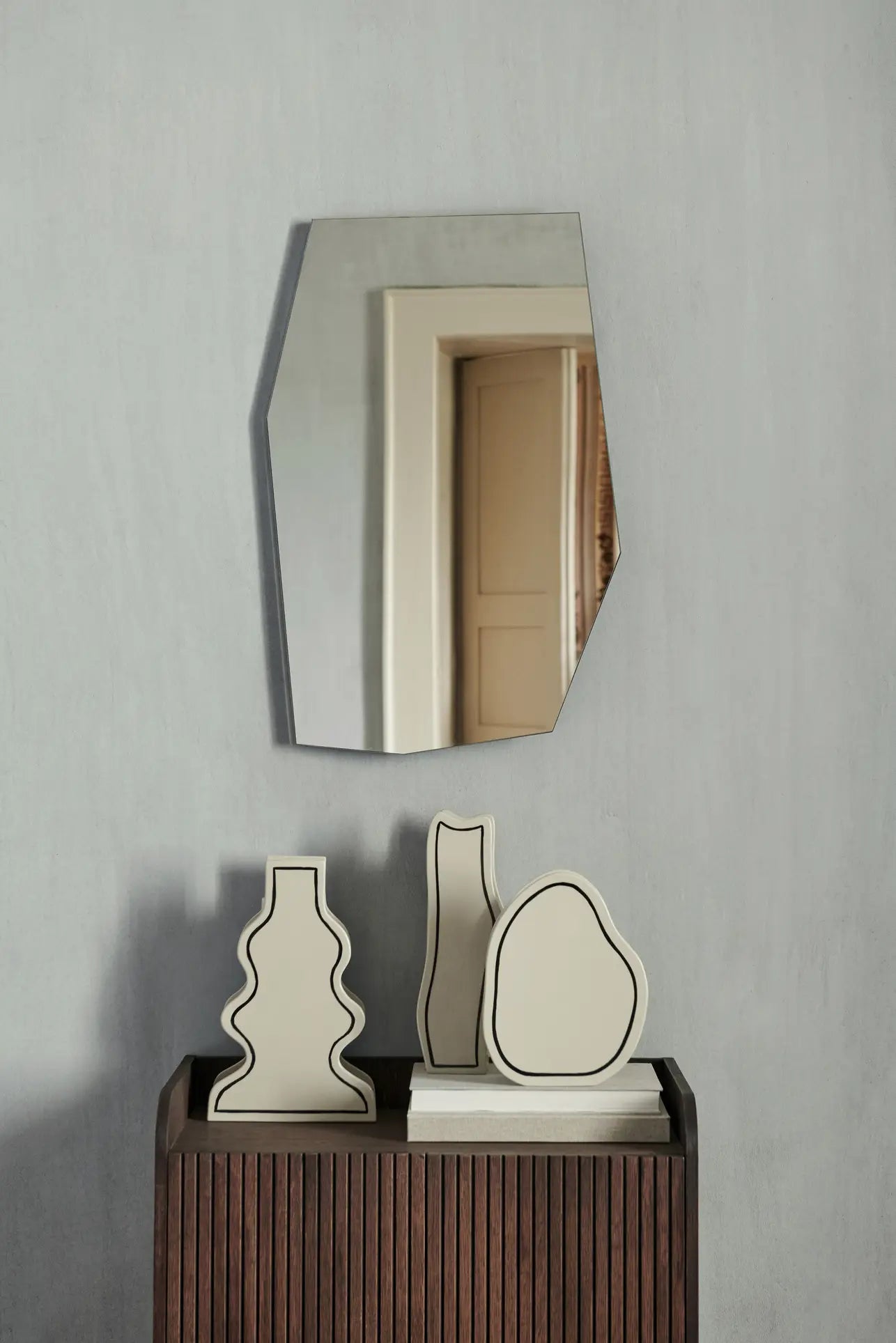 A geometric mirror hangs on a gray wall above a wooden cabinet displaying three ferm LIVING Paste Vase, Curvy ceramics in beige, each with black outlines and abstract shapes. The mirror reflects a doorway in the background.