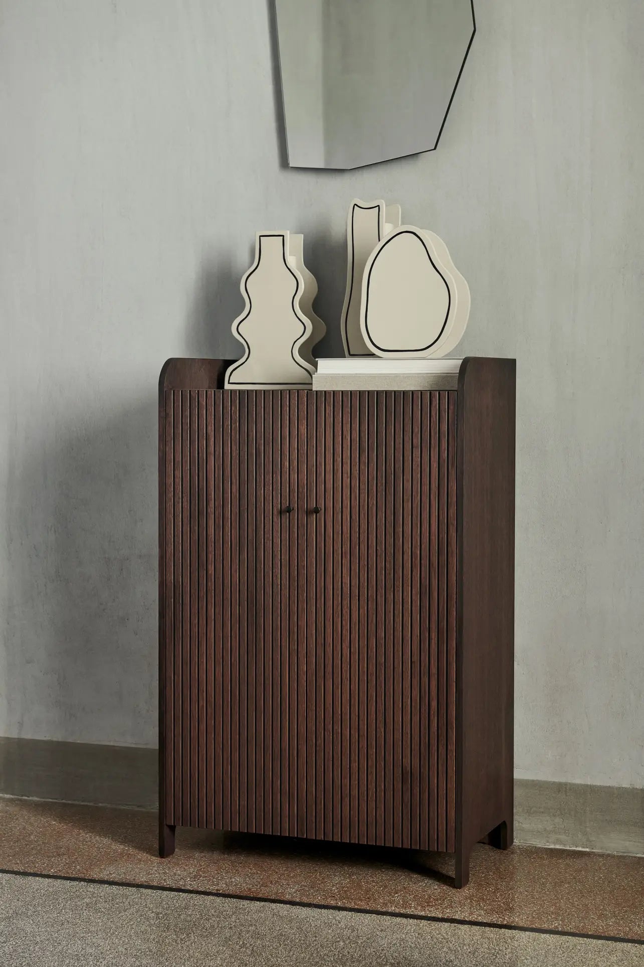 Against a gray wall stands a dark wooden cabinet with vertical grooves. On top are three cream-colored ferm LIVING Paste Vases, Curvy, crafted from ceramic sheets. Above, a geometric mirror highlights their unique design.