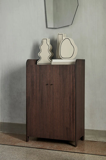 Against a gray wall stands a dark wooden cabinet with vertical grooves. On top are three cream-colored ferm LIVING Paste Vases, Curvy, crafted from ceramic sheets. Above, a geometric mirror highlights their unique design.