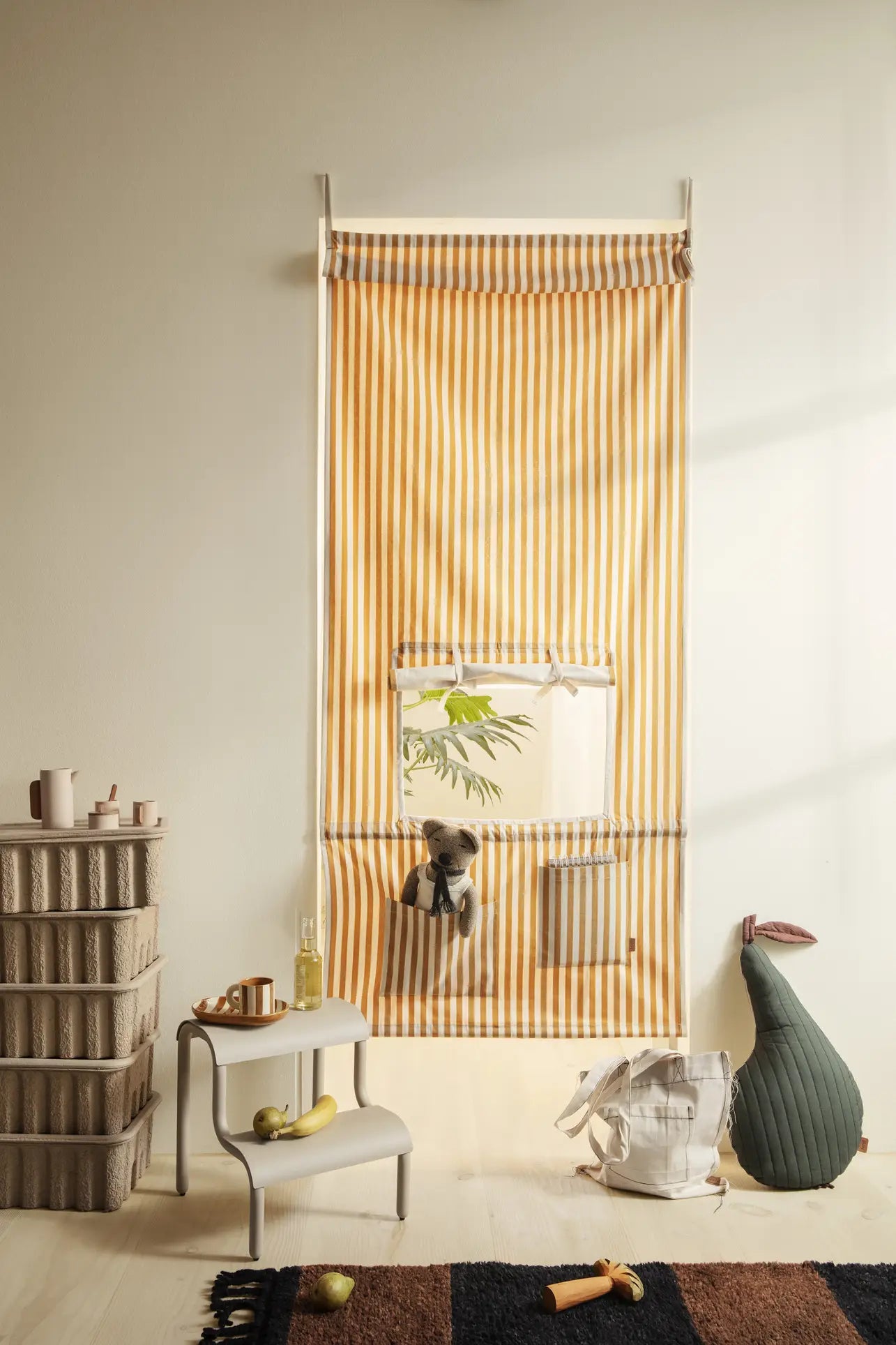 A playroom with a yellow and white striped fabric door cover featuring a window and a small gray toy. Nearby are shelves, a table with banana and bottle, rug, the ferm LIVING Pear Quilted Cushion (GOTS certified), and a white bag.