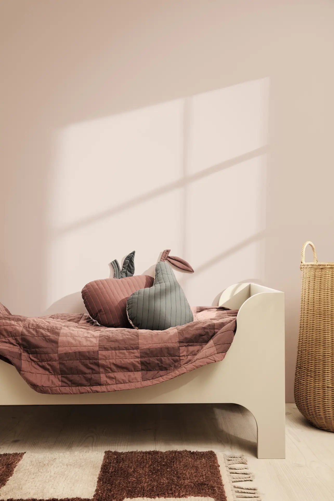 A minimalist kids’ room with a cream bed, mauve checkered organic cotton bedding, bunny-shaped pillows, a ferm LIVING Pear Quilted Cushion, a wicker basket, and sunlight casting shadows on the pale pink wall.