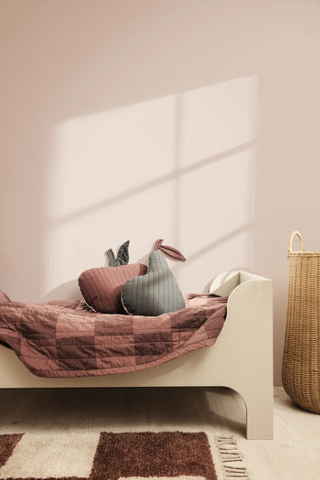 A minimalist kids’ room with a cream bed, mauve checkered organic cotton bedding, bunny-shaped pillows, a ferm LIVING Pear Quilted Cushion, a wicker basket, and sunlight casting shadows on the pale pink wall.