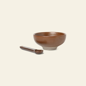The ferm LIVING Petite Salt Set features a textured brown ceramic bowl and matching spoon, elegantly displayed on a light beige background—a stylish and essential addition to any kitchen.