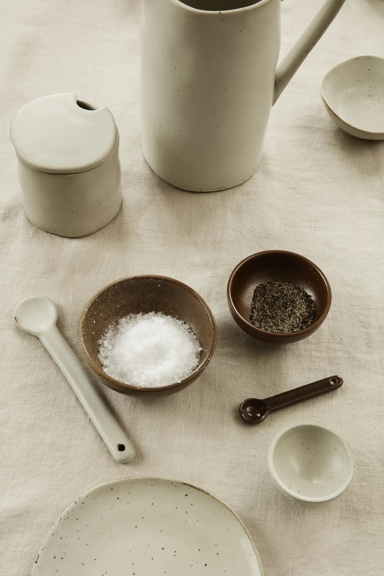 Minimalist ceramic dishes and utensils by ferm LIVING on a light cloth, featuring the Petite Salt Set, pitcher, small bowls with salt and pepper, a lidded jar, plate, and spoons. Neutral tones highlight each textured kitchen essential.