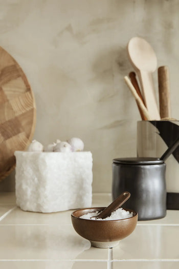The ferm LIVING Petite Salt Set, featuring a ceramic bowl filled with salt and a small wooden spoon, is displayed on a tiled kitchen counter beside a black jar, utensil holder with wooden spoons, and a white container holding garlic bulbs.