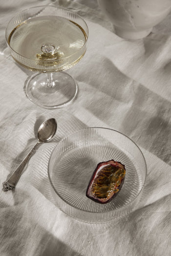 A mouth-blown coupe filled with a clear drink sits on a white textured cloth next to the ferm LIVING Ripple Serving Bowls, Set of 4, one holding half a passion fruit and a silver spoon.