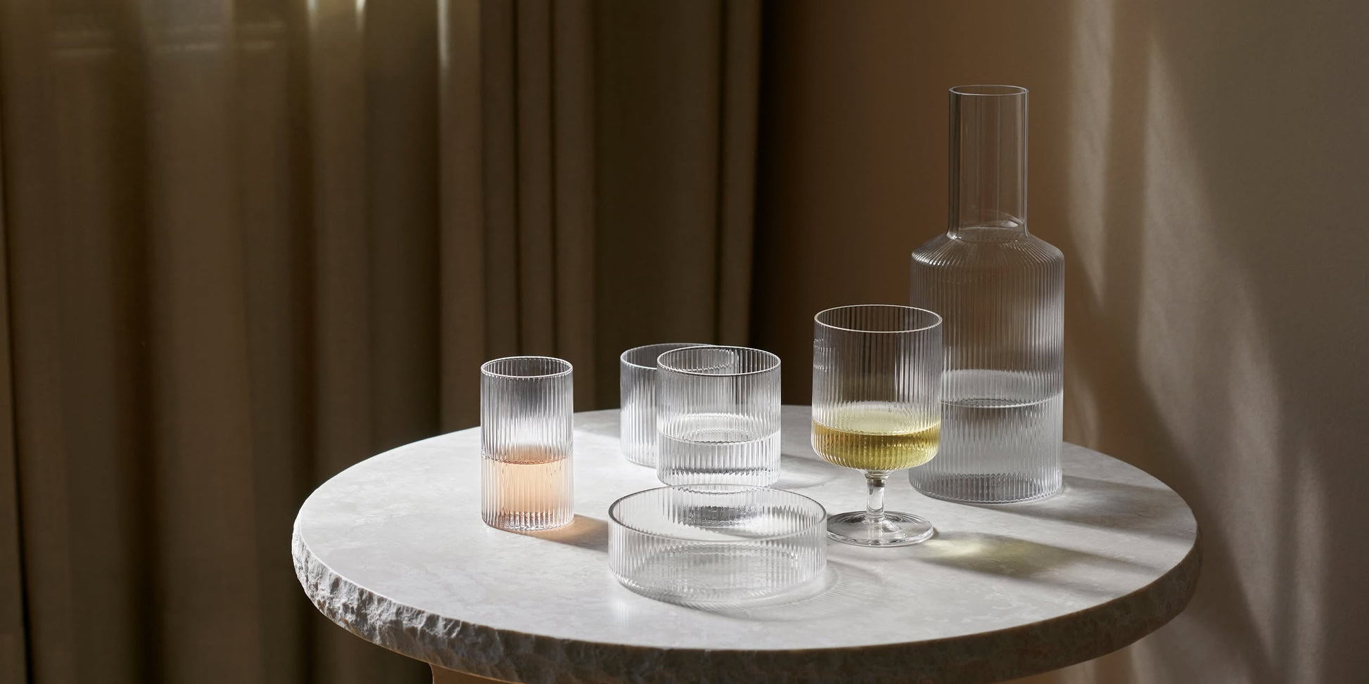 A round marble table with a textured surface displays a set of four ferm LIVING Ripple Serving Bowls, their ribbed glass catching sunlight as it streams softly through nearby curtains.