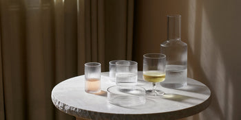 A round marble table with a textured surface displays a set of four ferm LIVING Ripple Serving Bowls, their ribbed glass catching sunlight as it streams softly through nearby curtains.