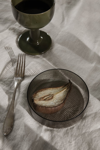 A sliced, dried pear sits on a ferm LIVING Ripple Serving Bowl beside a silver fork and a dark glass goblet, all elegantly arranged on a textured surface.