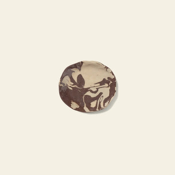 A round cookie with marbled beige and brown swirls rests on an off-white surface, reflecting the Nerikomi-inspired patterns found in the ferm LIVING Ryu Platter, 18.5cm by ferm LIVING.