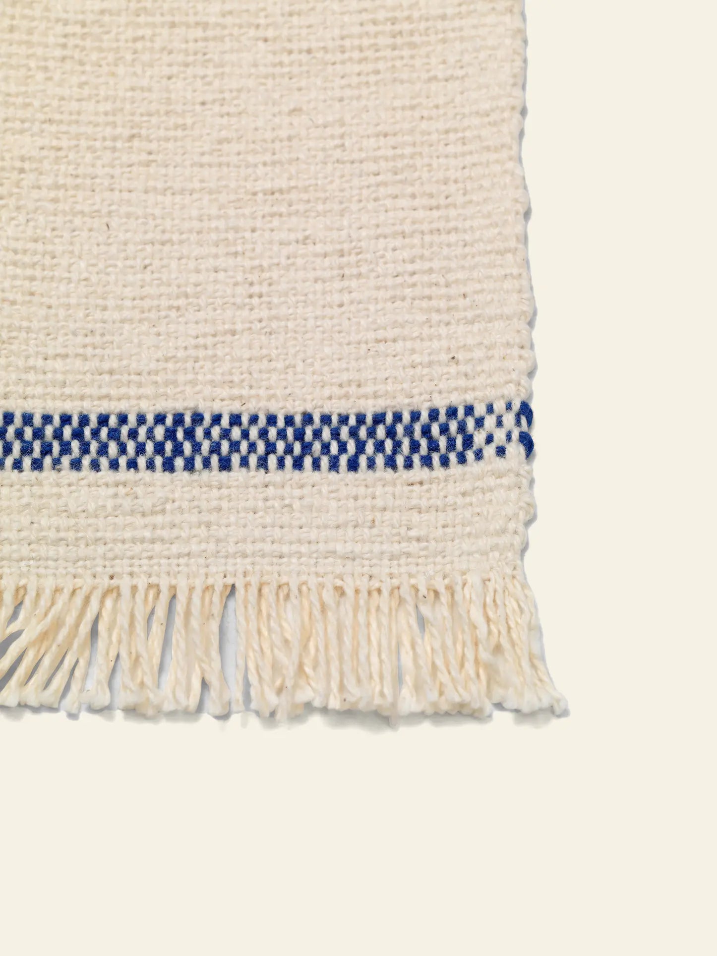 A close-up of ferm LIVING Savor Coasters, Set of 4, shows woven cream fabric with fringed edges and a horizontal blue checkered stripe near the bottom on a plain light background.