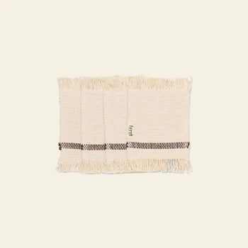 A set of four rectangular, cream-colored woven Savor Coasters by ferm LIVING, each with fringe edges and a brown checkered stripe near one side, shown on a plain light background.