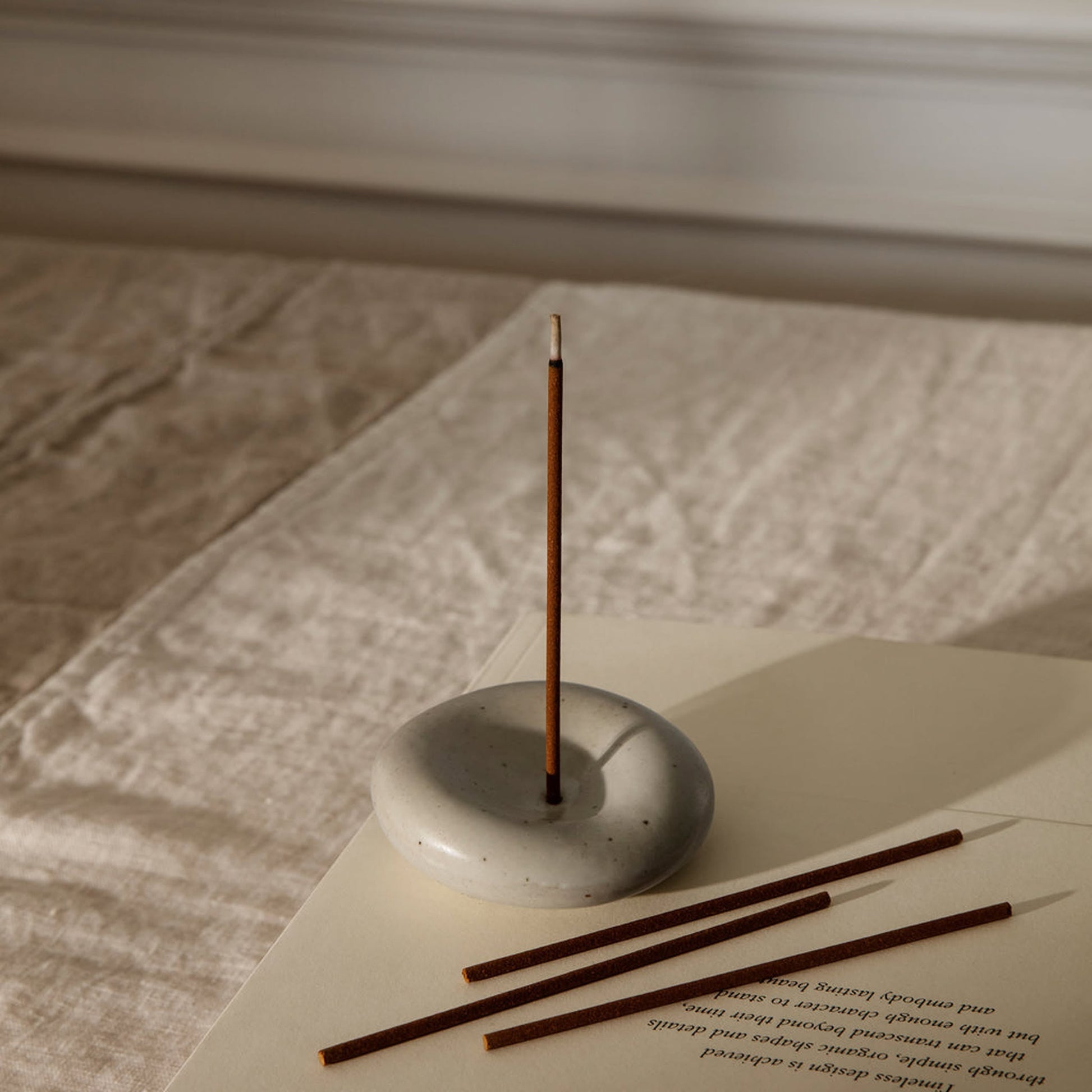 The ferm LIVING Sense Incense Holder Gift Set features a single sandalwood incense stick burning in its smooth, round holder on a beige cloth. Nearby, three unlit sticks and an open book create a calming scene—perfect for gifting by ferm LIVING.