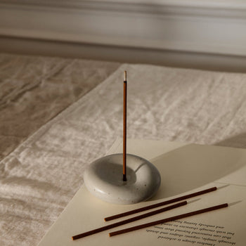 The ferm LIVING Sense Incense Holder Gift Set features a single sandalwood incense stick burning in its smooth, round holder on a beige cloth. Nearby, three unlit sticks and an open book create a calming scene—perfect for gifting by ferm LIVING.