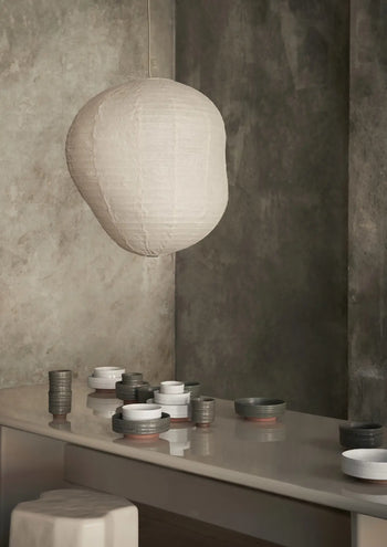 A soft, white paper lantern hangs above a glossy table set with neatly arranged ferm LIVING Serena Bowls (12.5cm) and cups in neutral tones, against a textured gray wall in a modern, serene interior.