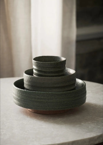 The ferm LIVING Serena Bowl (12.5cm) by ferm LIVING, in dark green ceramic with a luxurious texture, is stacked with matching bowls on a light round table, soft neutral curtains in the background.