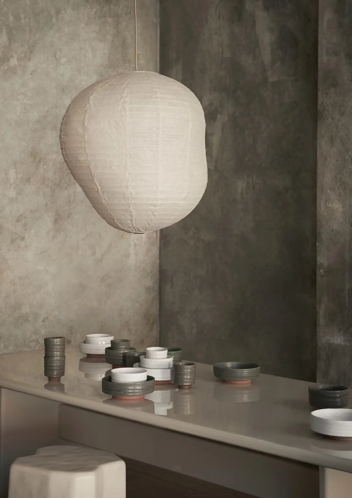 A modern, minimalist space features a large white lantern over a glossy surface displaying the ferm LIVING Serena Bowl, 12.5cm, along with other neutral-toned ceramics against a textured gray wall.