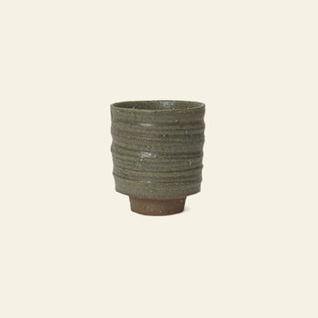 The ferm LIVING Serena Cup by ferm LIVING is a handcrafted clay cup with a ribbed, textured surface in earthy green and a reactive glaze, featuring a slightly raised base against a plain light background.
