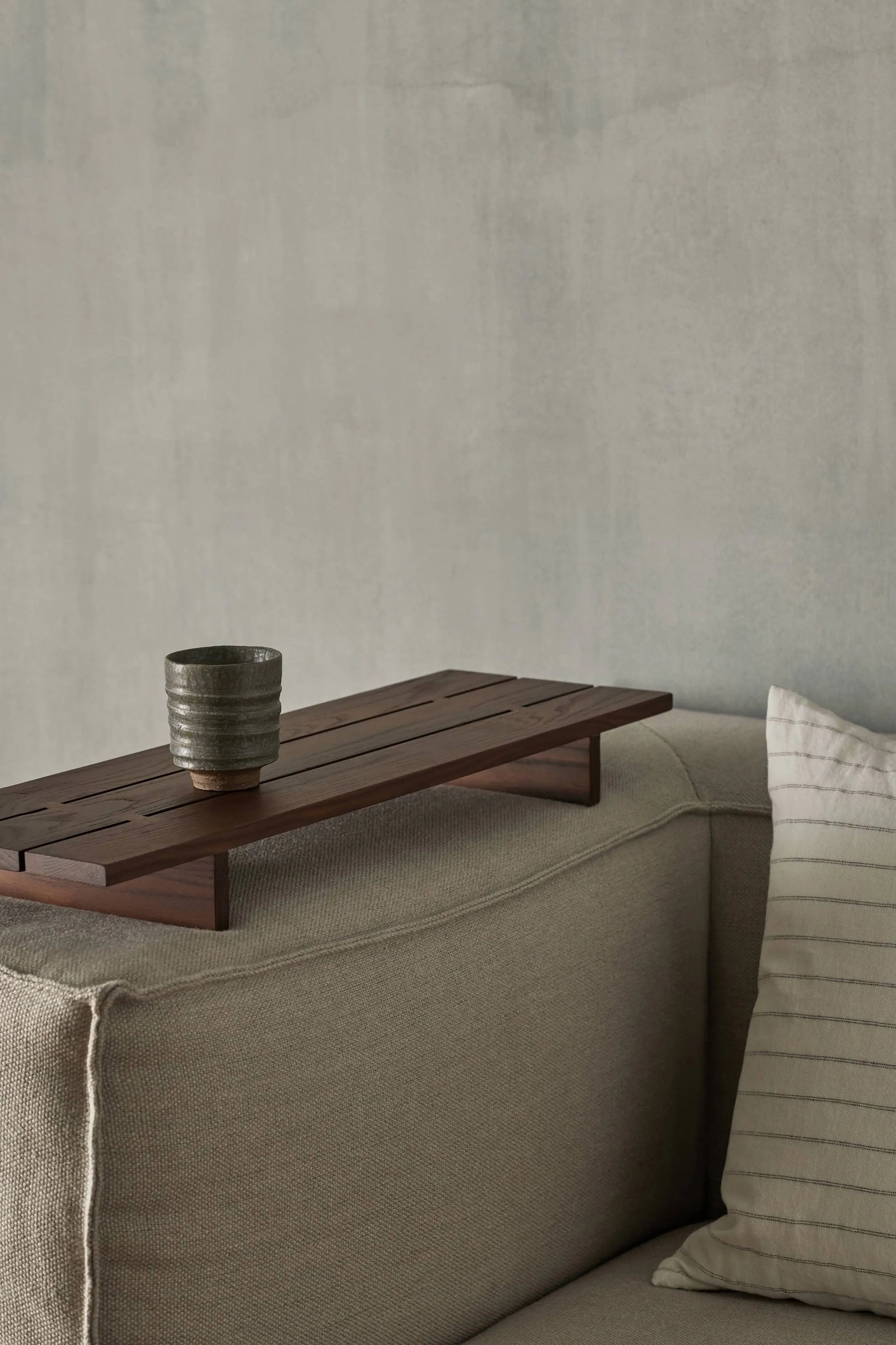 The ferm LIVING Serena Cup with a reactive glaze rests on a wooden tray atop a beige sofa’s armrest, beside a light striped cushion. A plain, light gray wall forms the background.